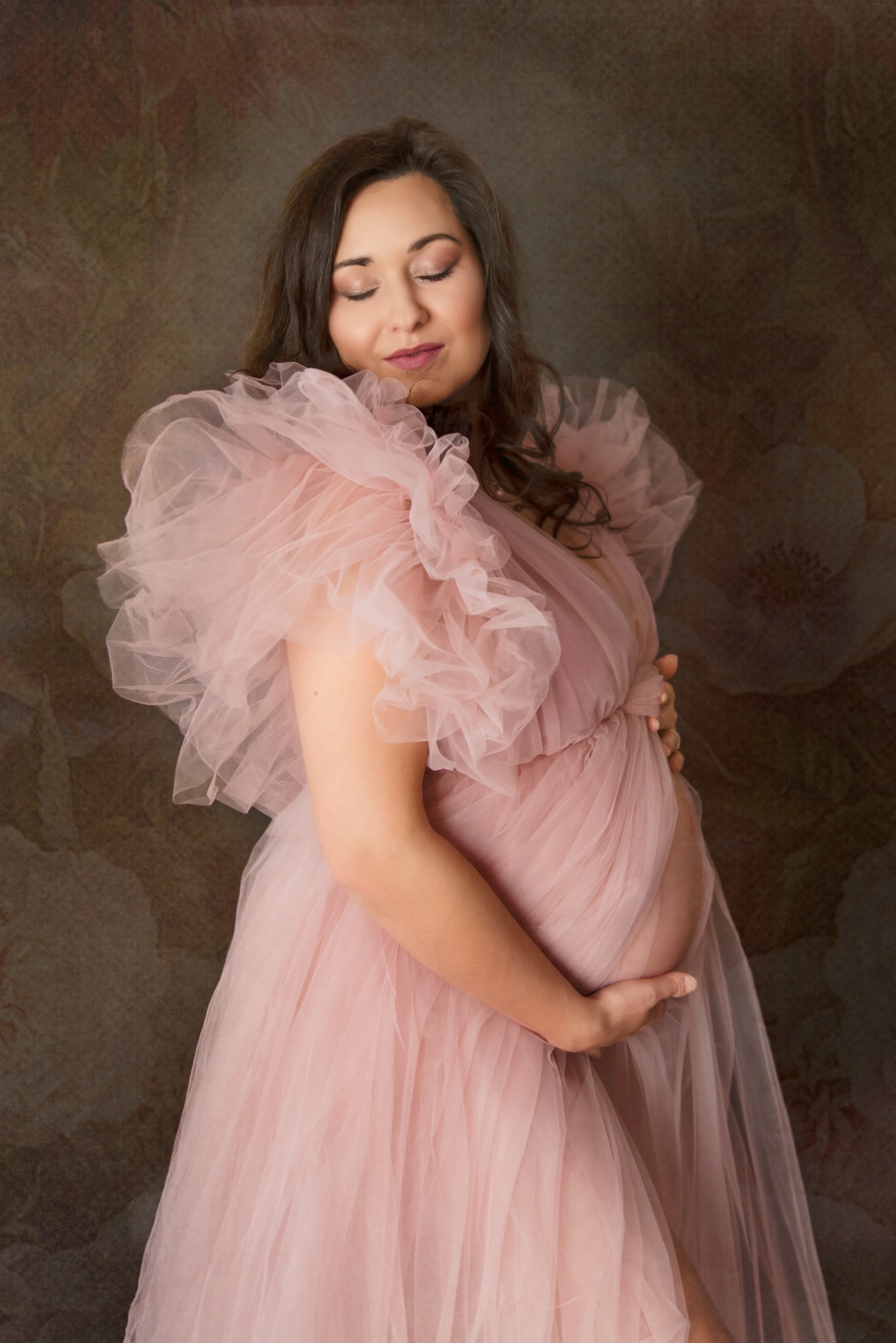 Kobieta w ciąży w pudrowo-różowej, tiulowej sukni, z zamkniętymi oczami, trzymająca dłonie na brzuchu na tle abstrakcyjnego tła w odcieniach brązu i różu.