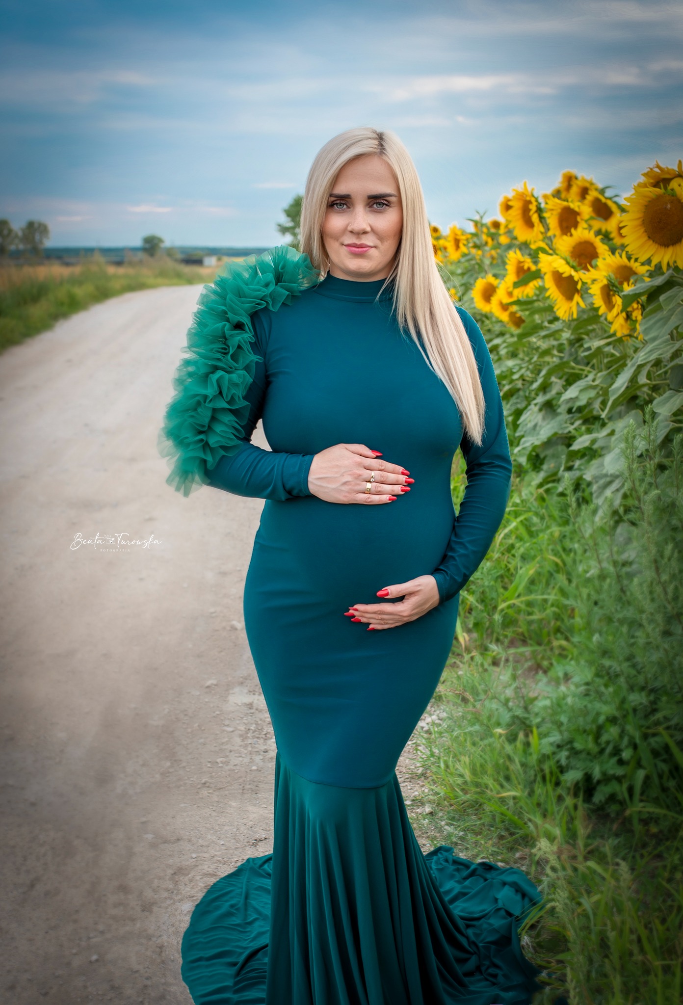 Kobieta w obcisłej, długiej, szmaragdowej sukni z falbaną na ramieniu, pozuje na polnej drodze z polem słoneczników w tle, trzymając dłonie na brzuchu.
