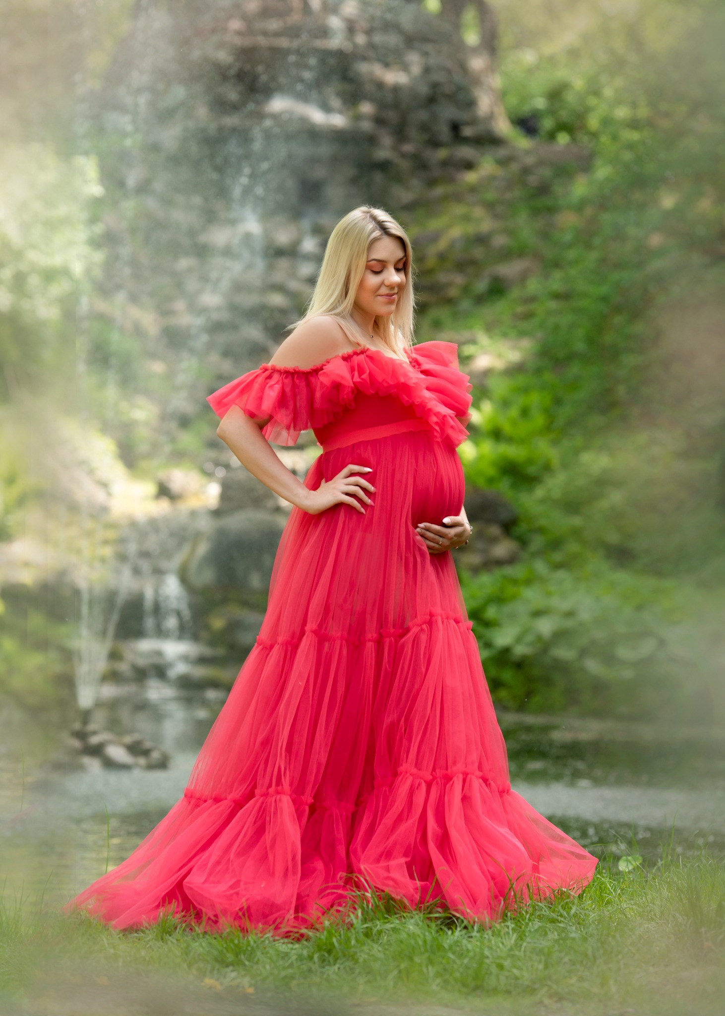 Kobieta w czerwonej, zwiewnej sukni pozuje z ręką na brzuchu na tle wodospadu i zieleni.