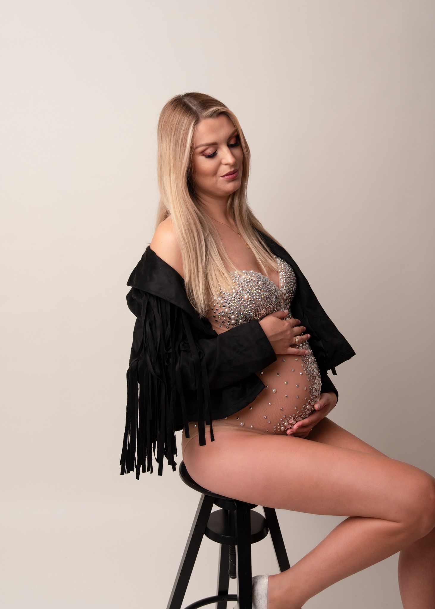 Kobieta w ciąży siedzi na stołku, ubrana w cielisty strój zdobiony perłami i czarną kurtkę z frędzlami, patrzy z uśmiechem na brzuch.