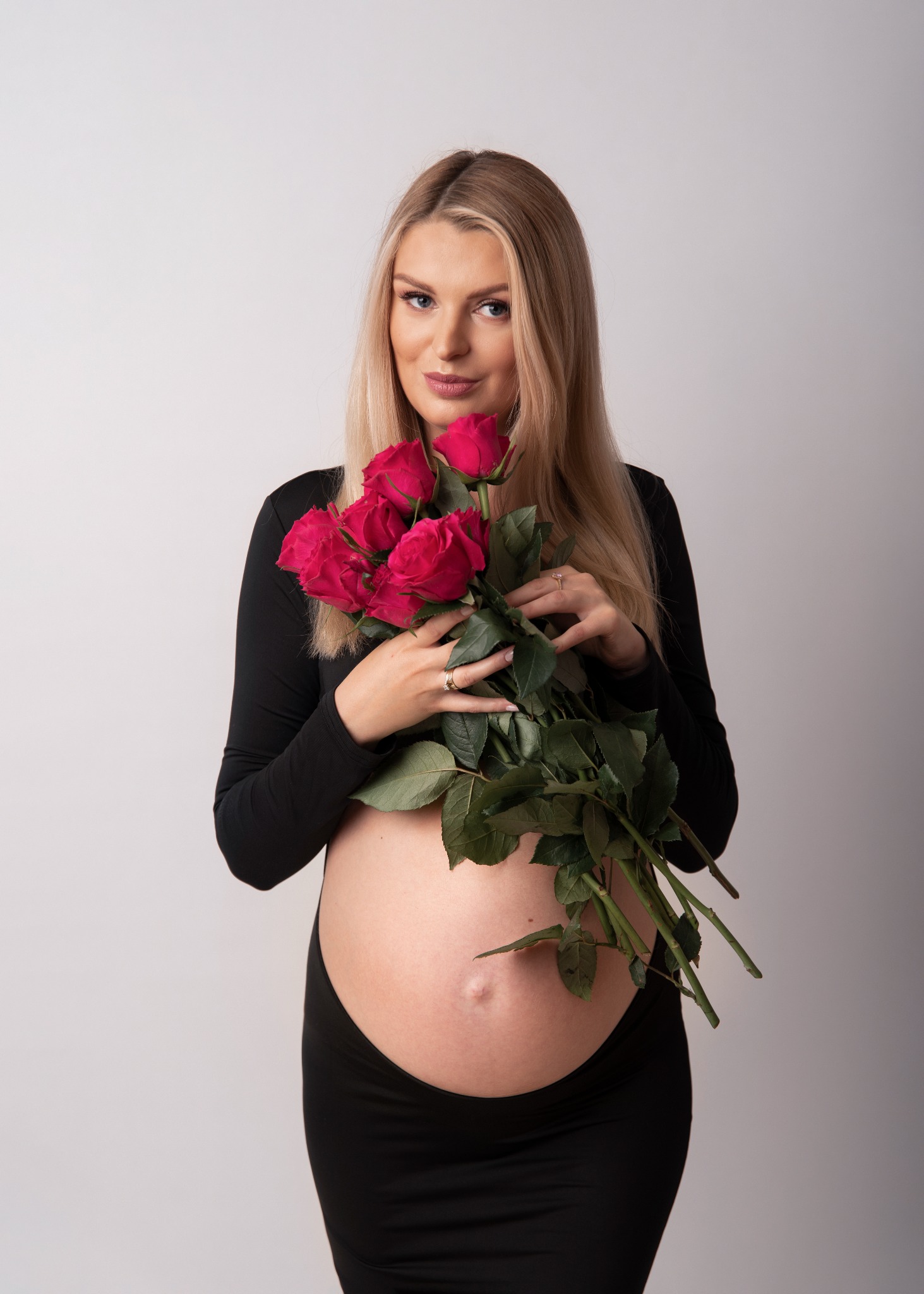 Portret kobiety w czarnej sukience ciążowej, trzymającej bukiet czerwonych róż na jasnym tle.