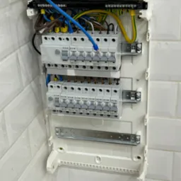 Otwarta skrzynka bezpiecznikowa z widocznymi bezpiecznikami EATON, przewodami w r&oacute;żnych kolorach (niebieski, brązowy, ż&oacute;łto-zielony) i listwami zaciskowymi, zamontowana na ścianie z białych płytek.