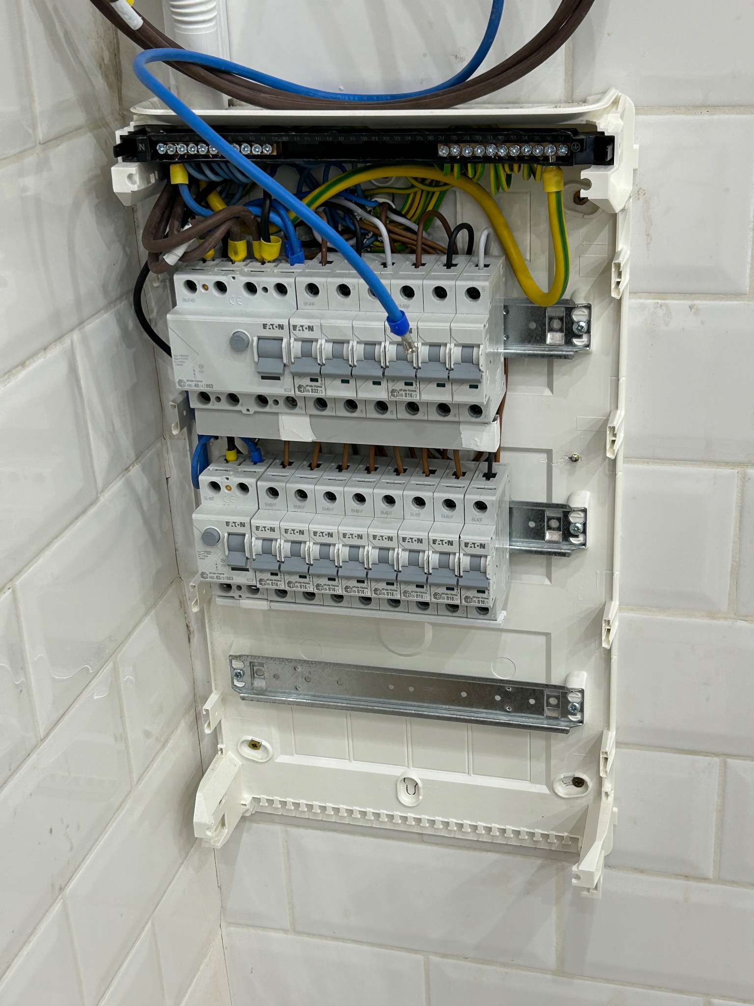 Otwarta skrzynka bezpiecznikowa z widocznymi bezpiecznikami EATON, przewodami w różnych kolorach (niebieski, brązowy, żółto-zielony) i listwami zaciskowymi, zamontowana na ścianie z białych płytek.