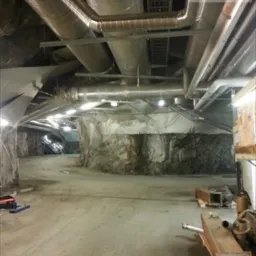 Wnętrze sztolni z widoczną instalacją wentylacyjną pod sufitem, obejmującą rury i kanały wentylacyjne, oraz częściowo zakryte ściany skalne.