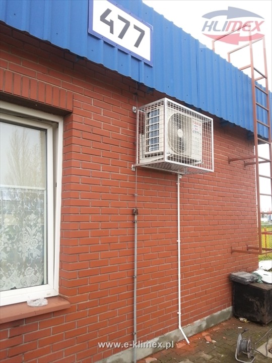 Zewnętrzna jednostka klimatyzacji w metalowej klatce ochronnej zamontowana na ceglanej ścianie budynku z numerem 477, widoczna drabina i logo firmy w prawym górnym rogu.
