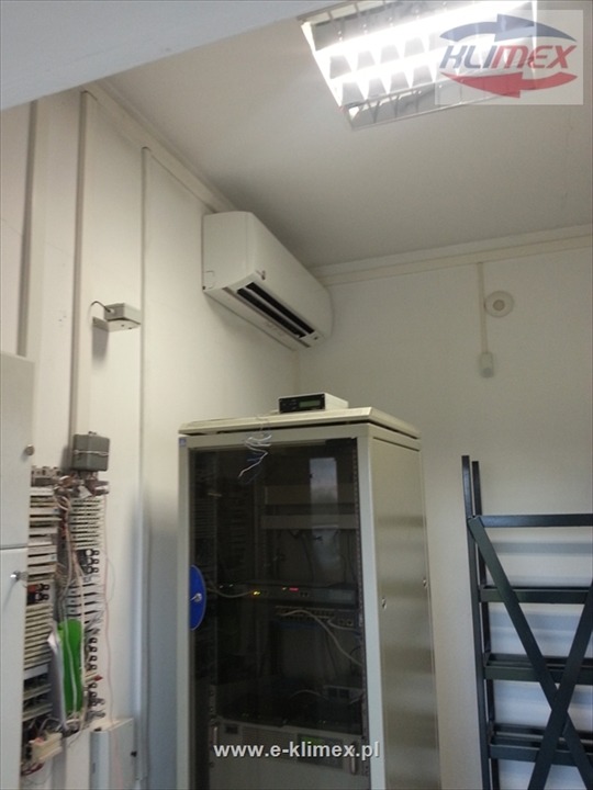 Biały klimatyzator ścienny zamontowany w pomieszczeniu serwerowni, widoczna szafa rack i okablowanie, logo firmy w rogu kadru.