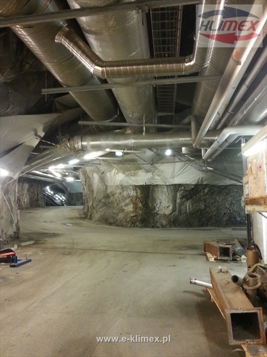 Wnętrze sztolni z widoczną instalacją wentylacyjną pod sufitem, obejmującą rury i kanały wentylacyjne, oraz częściowo zakryte ściany skalne.