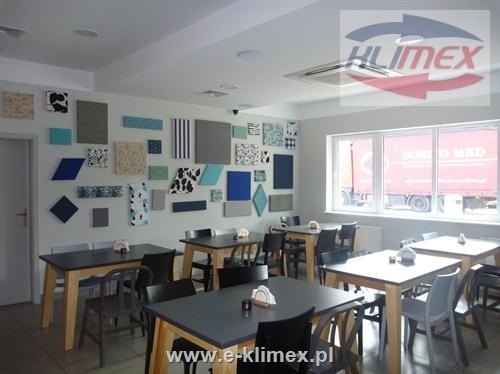 Wnętrze lokalu gastronomicznego z zamontowaną kasetonową jednostką klimatyzacyjną w suficie, widoczny dekor ścienny z paneli akustycznych oraz logo firmy na urządzeniu.