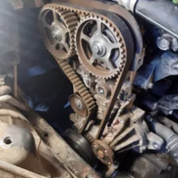 Silnik samochodowy z widocznym paskiem rozrządu i zębatkami podczas naprawy, zabrudzone elementy silnika, oświetlenie warsztatowe.