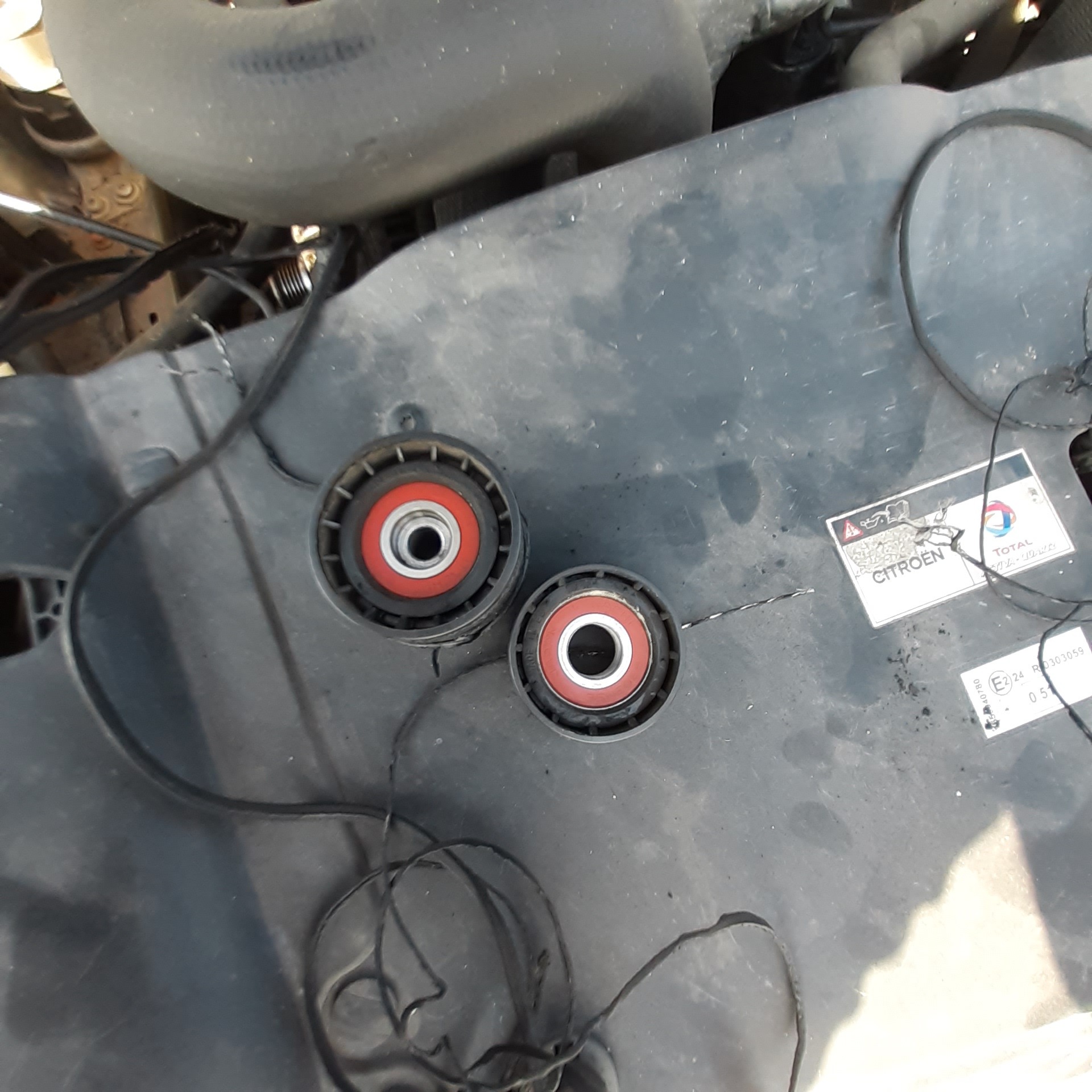 Dwa zużyte napinacze paska klinowego z czerwonymi łożyskami, leżące na brudnej, plastikowej osłonie silnika Citroena, widoczne przewody.