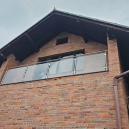 Szklana balustrada balkonowa z metalowymi słupkami, zamontowana na ceglanym budynku z widocznym dachem i rynną z flagą.