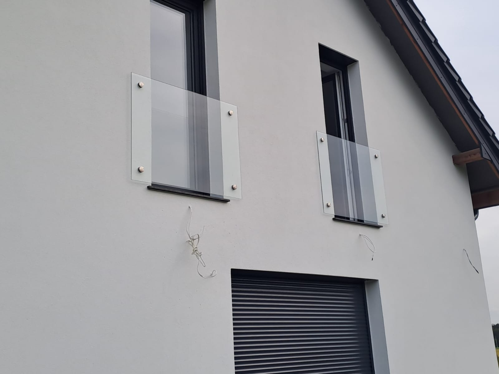 Nowoczesny dom z białym tynkiem, prezentujący szklane barierki balkonowe zamontowane przy oknach, z widocznymi śladami po instalacji w postaci zwisających przewodów.