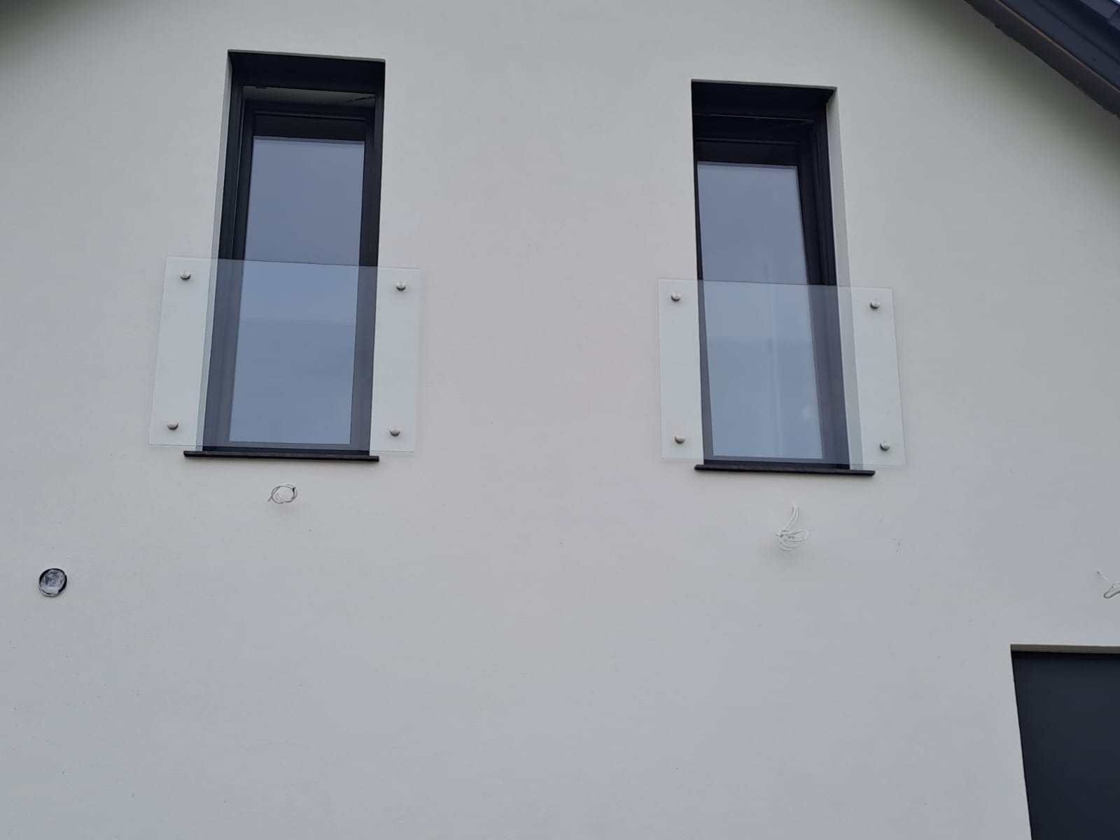Dwa okna na białej elewacji zabezpieczone szklanymi panelami mocowanymi punktowo, widoczne przewody instalacji elektrycznej