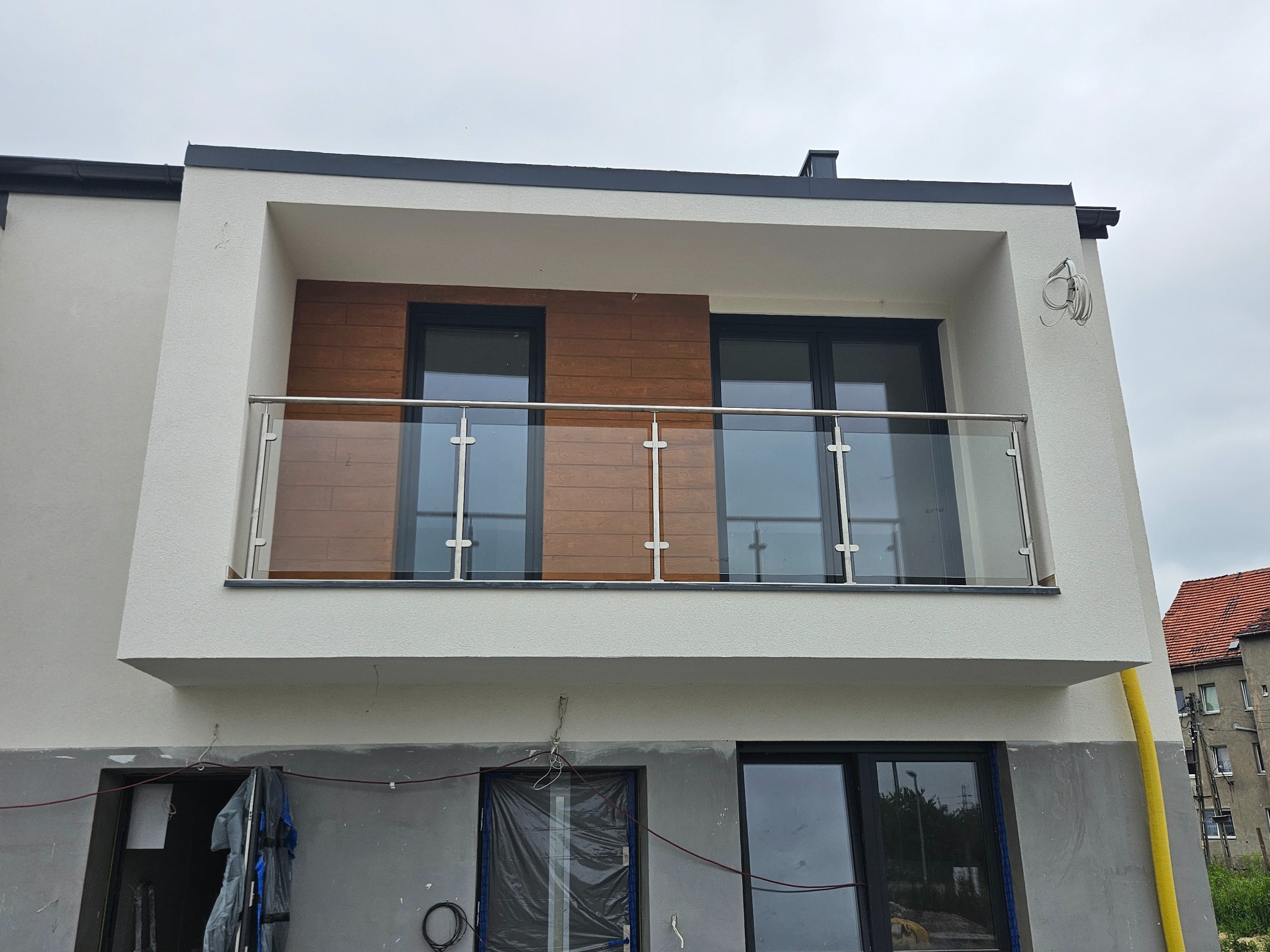 Nowoczesny balkon z przeszkloną balustradą, drewnianym wykończeniem ściany i widocznymi przewodami elektrycznymi na elewacji budynku w trakcie wykończenia.