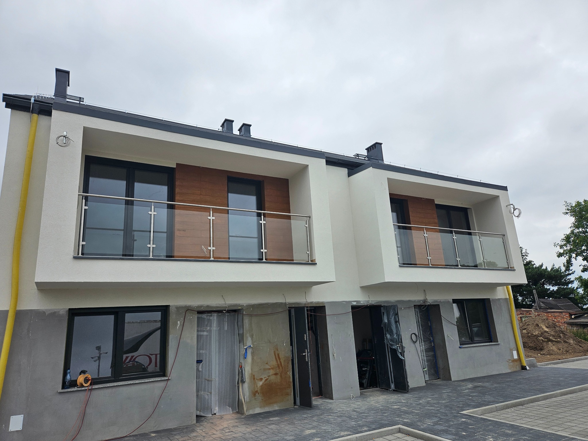 Nowoczesny dwupiętrowy budynek w trakcie wykończenia z balkonami zabezpieczonymi balustradami szklanymi, widoczne niedokończone elewacje i prowizoryczne zabezpieczenia wejść.