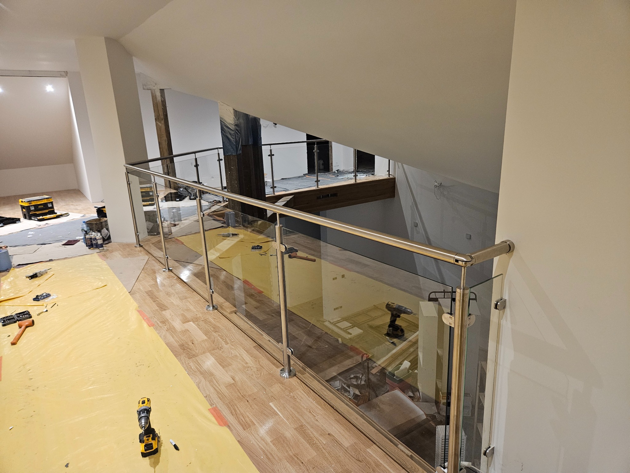 Montaż balustrady ze szkła i stali nierdzewnej na antresoli, widoczne narzędzia i zabezpieczenia podłogi.