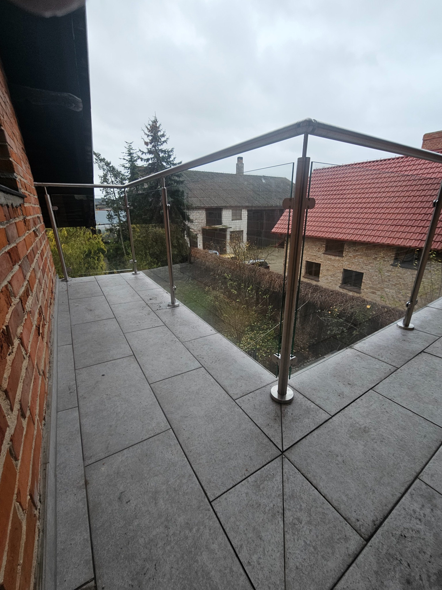 Balkon z nowoczesną, szklaną balustradą na tle zabudowań mieszkalnych w pochmurny dzień.