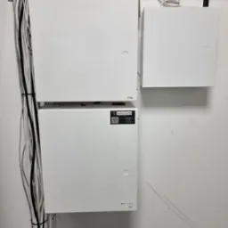 Trzy białe skrzynki elektryczne zamontowane na ścianie, z widocznymi kablami przytwierdzonymi do ściany po lewej stronie. Na skrzynkach widoczne etykiety i logo 'GRADE SYSTEM'.