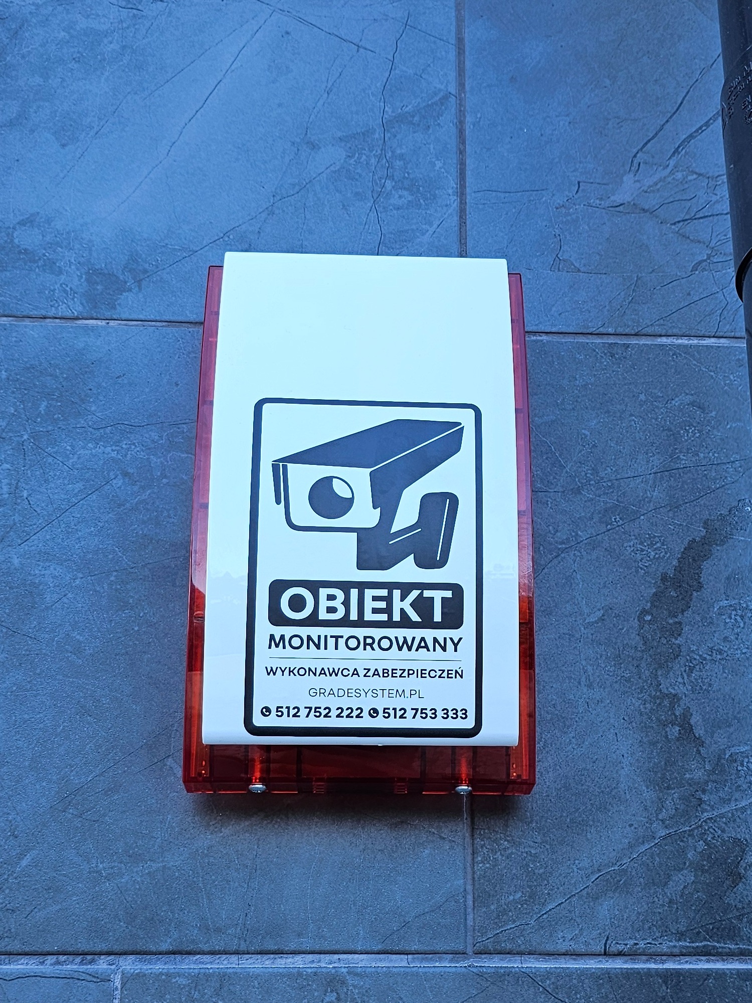 Tabliczka informacyjna z symbolem kamery CCTV i napisem 'Obiekt monitorowany', umieszczona na szarej, teksturowanej ścianie zewnętrznej budynku. Widoczne dane kontaktowe wykonawcy zabezpieczeń.