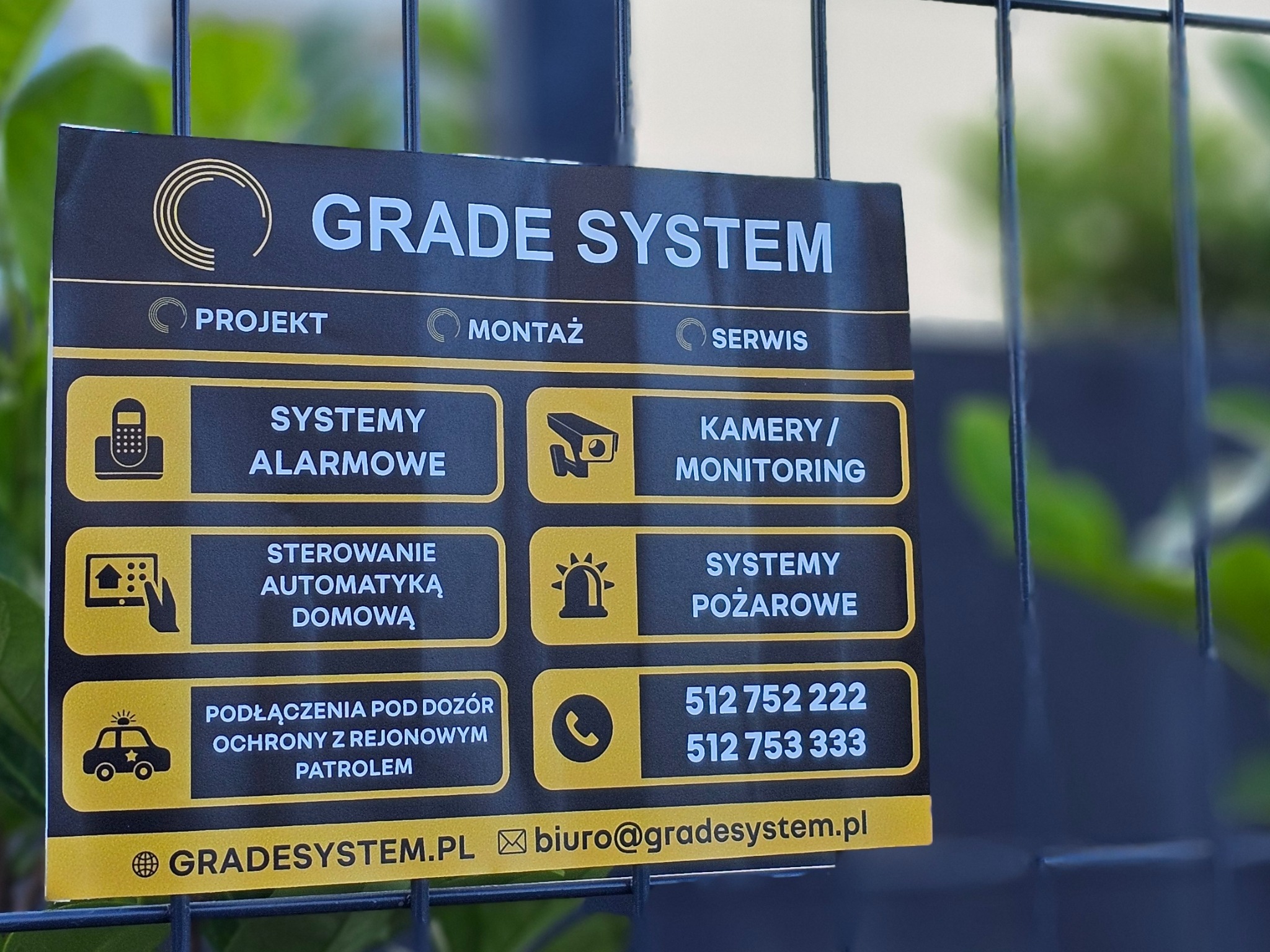 Tabliczka informacyjna firmy Grade System, zamocowana na metalowym ogrodzeniu, oferującej projekt, montaż i serwis systemów alarmowych, kamer/monitoringu, sterowania automatyką domową, systemów...