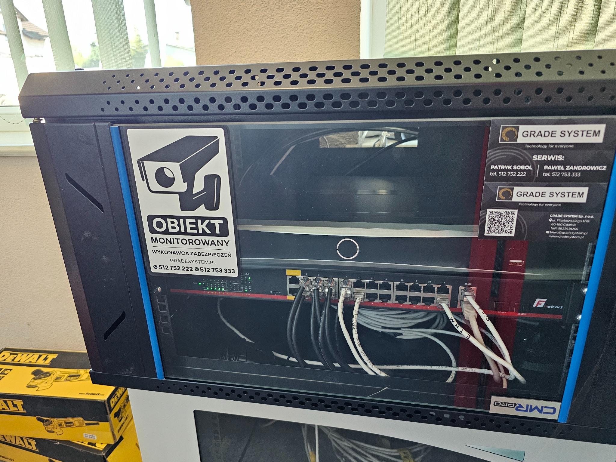 Szafa rack z zainstalowanym switchem sieciowym, okablowaniem oraz naklejką informującą o obiekcie monitorowanym i danych kontaktowych firmy Grade System, widoczne opakowania po elektronarzędziach...