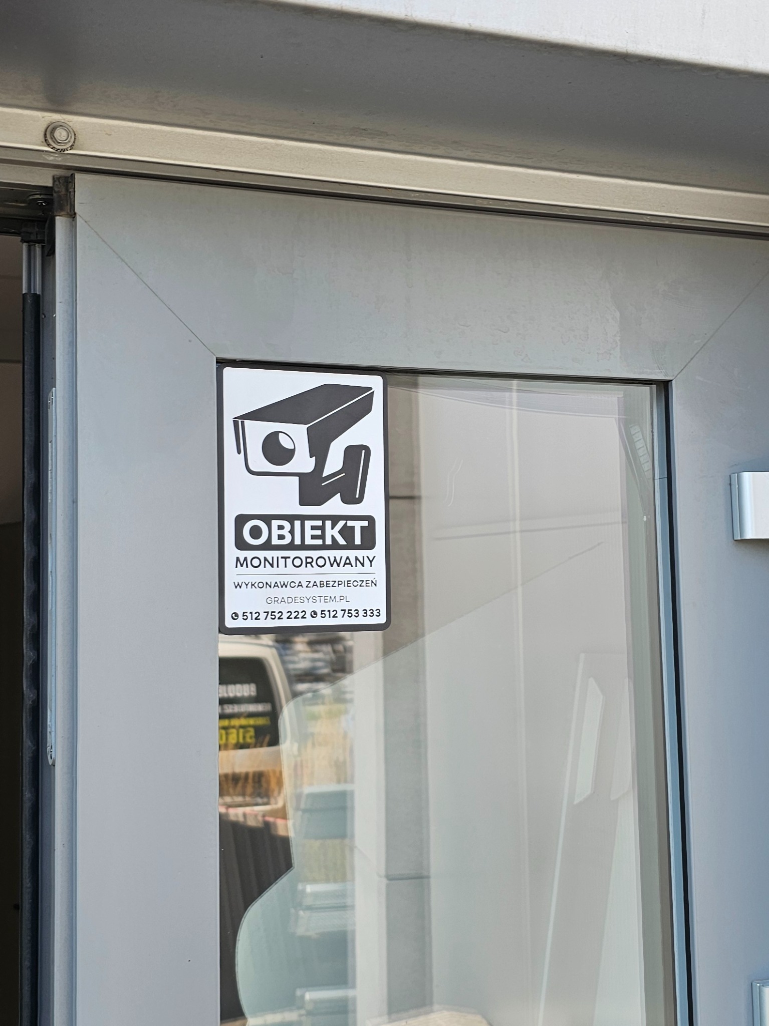 Naklejka informacyjna na szklanych drzwiach z symbolem kamery i napisem 'Obiekt monitorowany', zawierająca dane kontaktowe firmy ochroniarskiej.