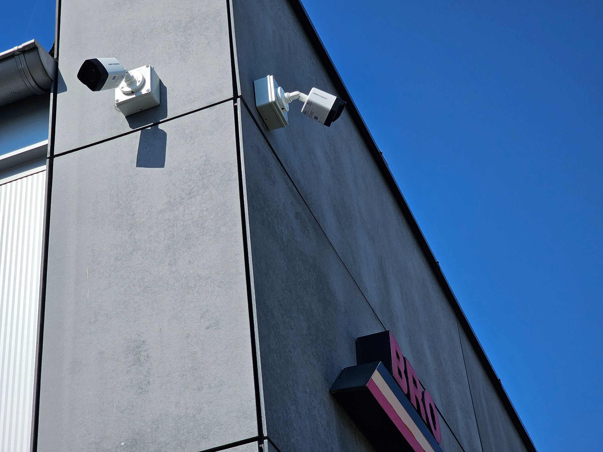 Dwie kamery monitoringu Hikvision zamontowane na szarej elewacji budynku z widocznym szyldem firmy na tle błękitnego nieba.
