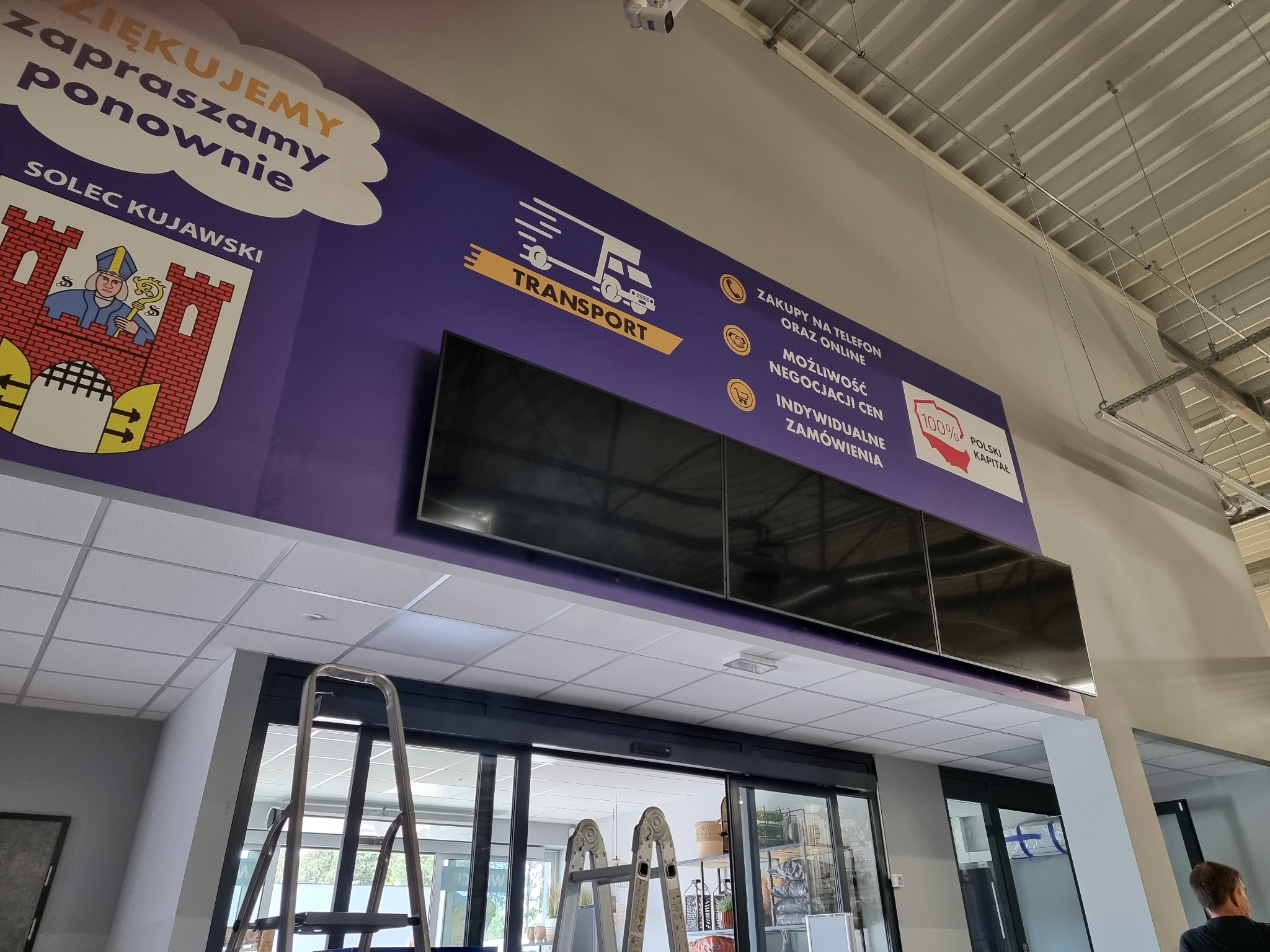 Instalacja monitorów w przestrzeni handlowej, widoczna drabina, logo 'Transport' i inne elementy graficzne na ścianie, w tle przesuwne drzwi wejściowe.