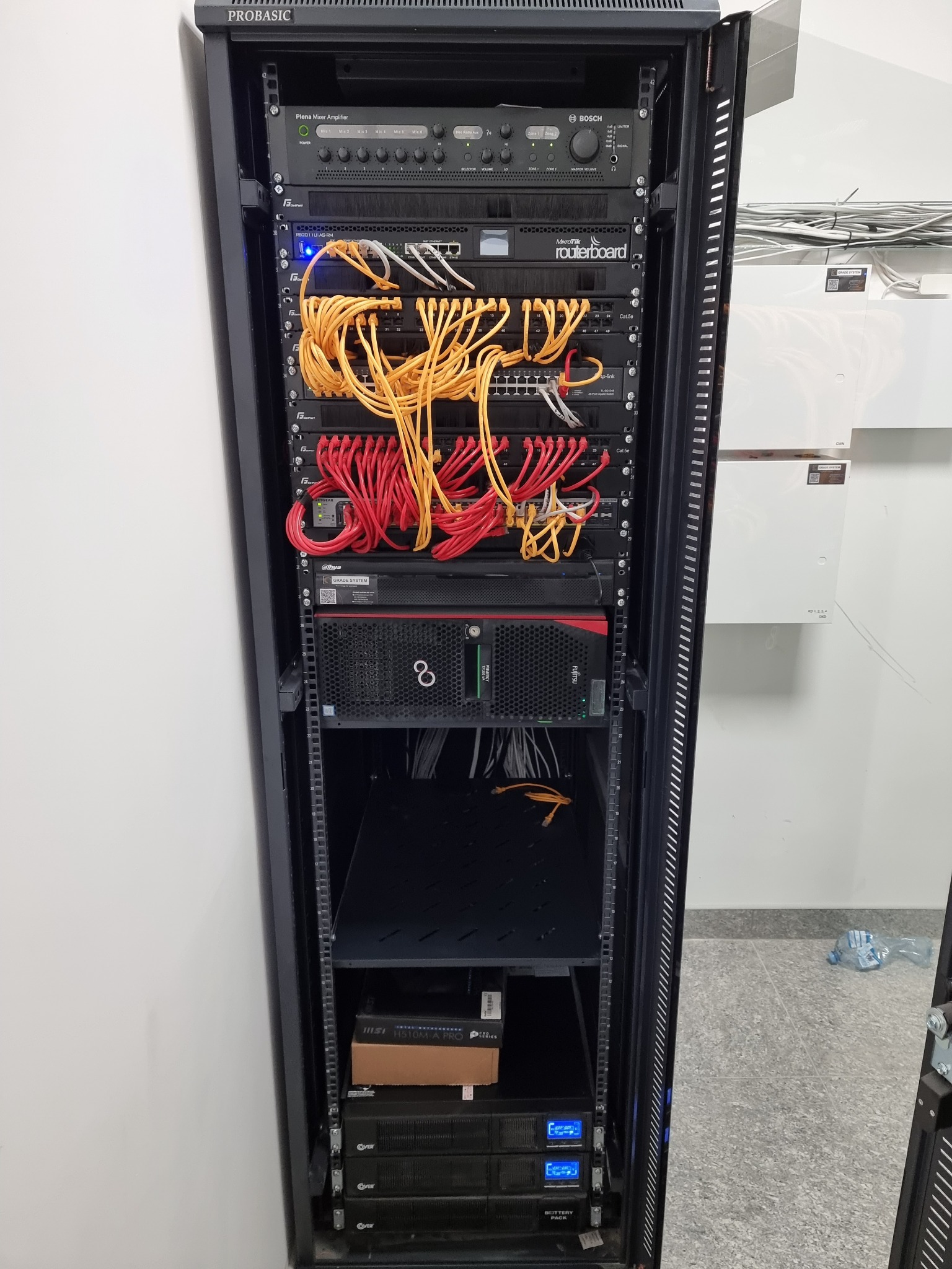 Szafa rack z zainstalowanymi urządzeniami sieciowymi: wzmacniacz Bosch Plena Mixer Amplifier, routerboard MikroTik, switche z okablowaniem Cat.5e w kolorach żółtym i czerwonym, serwer Fujitsu...