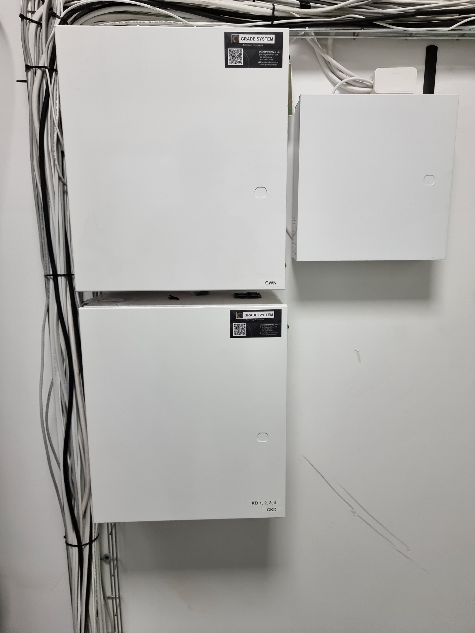 Trzy białe skrzynki elektryczne zamontowane na ścianie, z widocznymi kablami przytwierdzonymi do ściany po lewej stronie. Na skrzynkach widoczne etykiety i logo 'GRADE SYSTEM'.