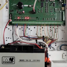 Centrala alarmowa z podłączonym akumulatorem i transformatorem, widoczne kable i elementy elektroniki, całość umieszczona w białej obudowie.