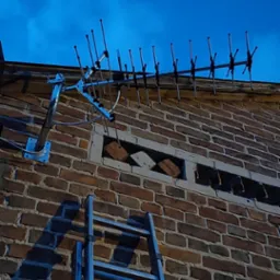 Antena telewizyjna zamontowana na ceglanym budynku, widoczna drabina oparta o ścianę, ujęcie z dołu na tle wieczornego nieba.