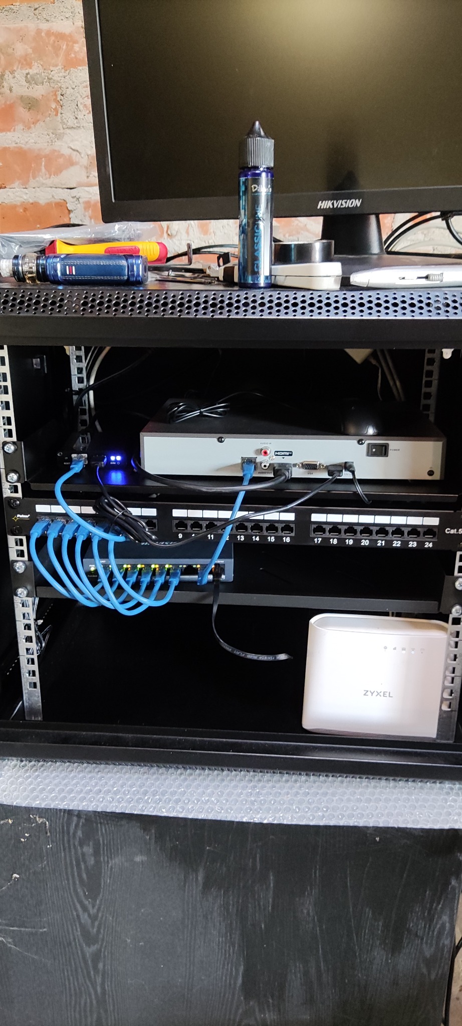 Zamknięta szafa rack z zamontowanym switchem sieciowym, patch panelem, routerem Zyxel i rejestratorem Hikvision. Na górze szafy leżą narzędzia i e-papieros.