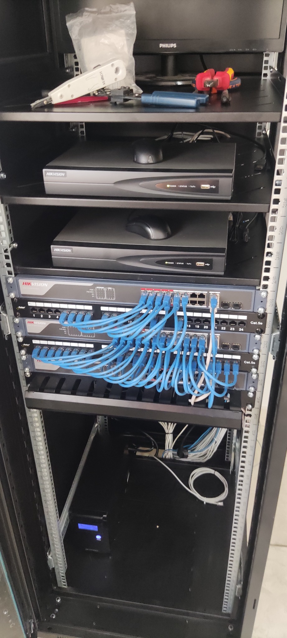 Szafa rack z zamontowanymi rejestratorami Hikvision, patch panelem z podłączonymi kablami sieciowymi Cat5e oraz narzędziami na górnej półce, takimi jak zaciskarka i śrubokręty.
