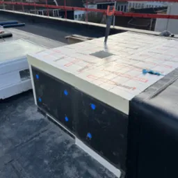 Izolacja termiczna dachu płaskiego z widoczną warstwą styropianu i papy, piła wbita w styropian, niebieskie kołki mocujące.