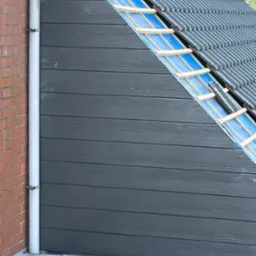 Fragment dachu z szarego panelu drewnopodobnego i częściowo ułożonej dachówki, widoczne elementy konstrukcyjne i rynna spustowa przy ceglanym murze.