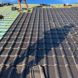 Układanie ciemnej dachówki na konstrukcji dachu z widocznym deskowaniem i membraną, częściowo zakończone, z narzędziami i stopami dekarza w kadrze, w słoneczny dzień.