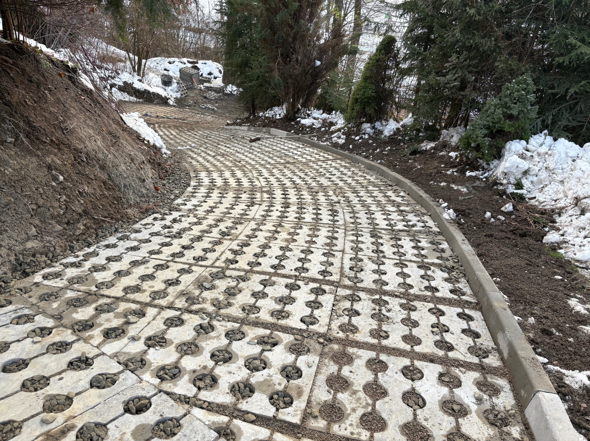 Ukończona ścieżka z ażurowych płyt betonowych wypełnionych drobnym żwirem, prowadząca pod górę w zimowym krajobrazie. Obrzeża ścieżki wykończone betonowym krawężnikiem.