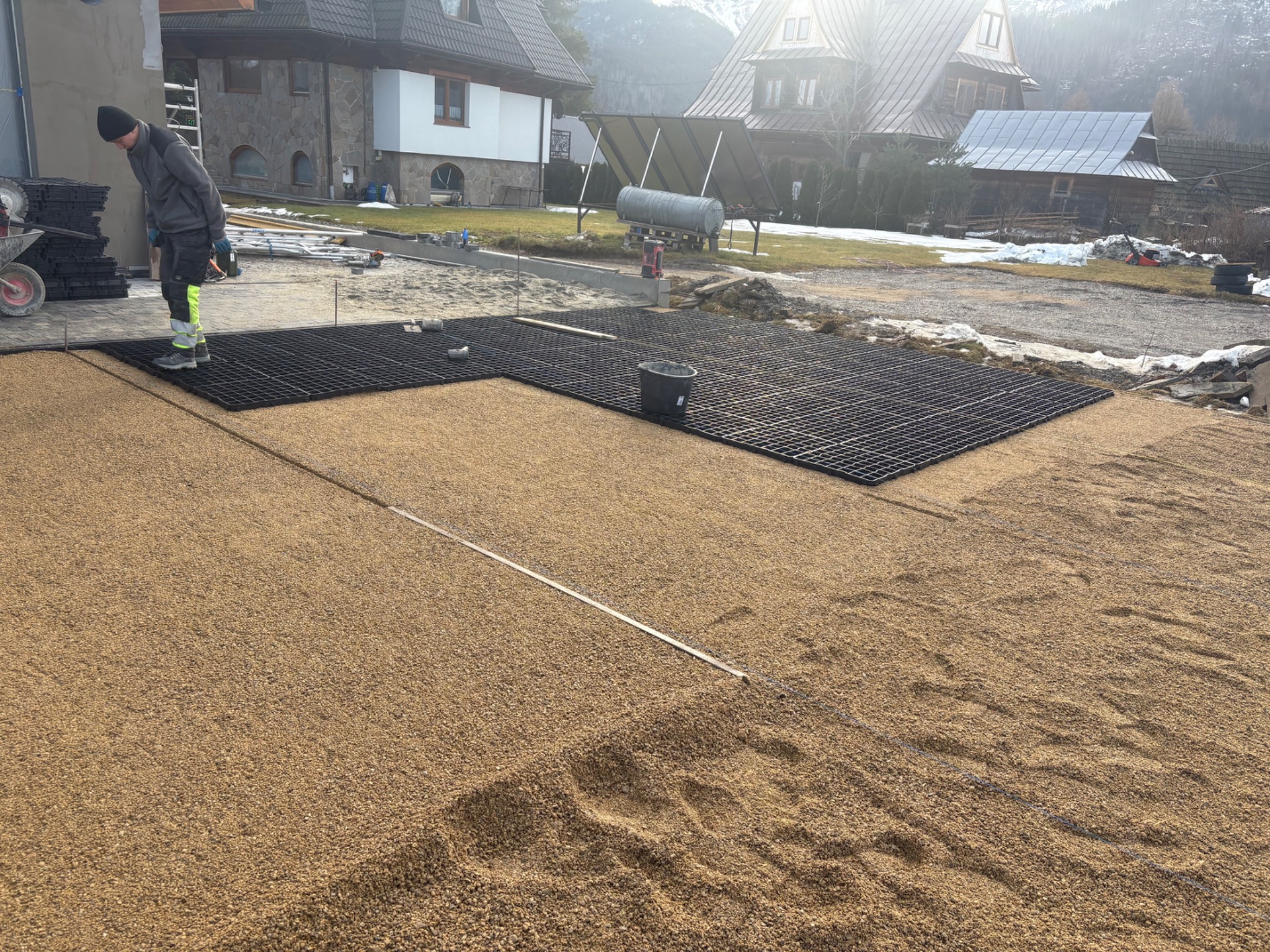 Montaż ażurowych płyt na podsypce piaskowej, widoczny pracownik w odblaskowych spodniach, w tle domy w stylu góralskim i góry. Etap przygotowania podłoża pod nawierzchnię.