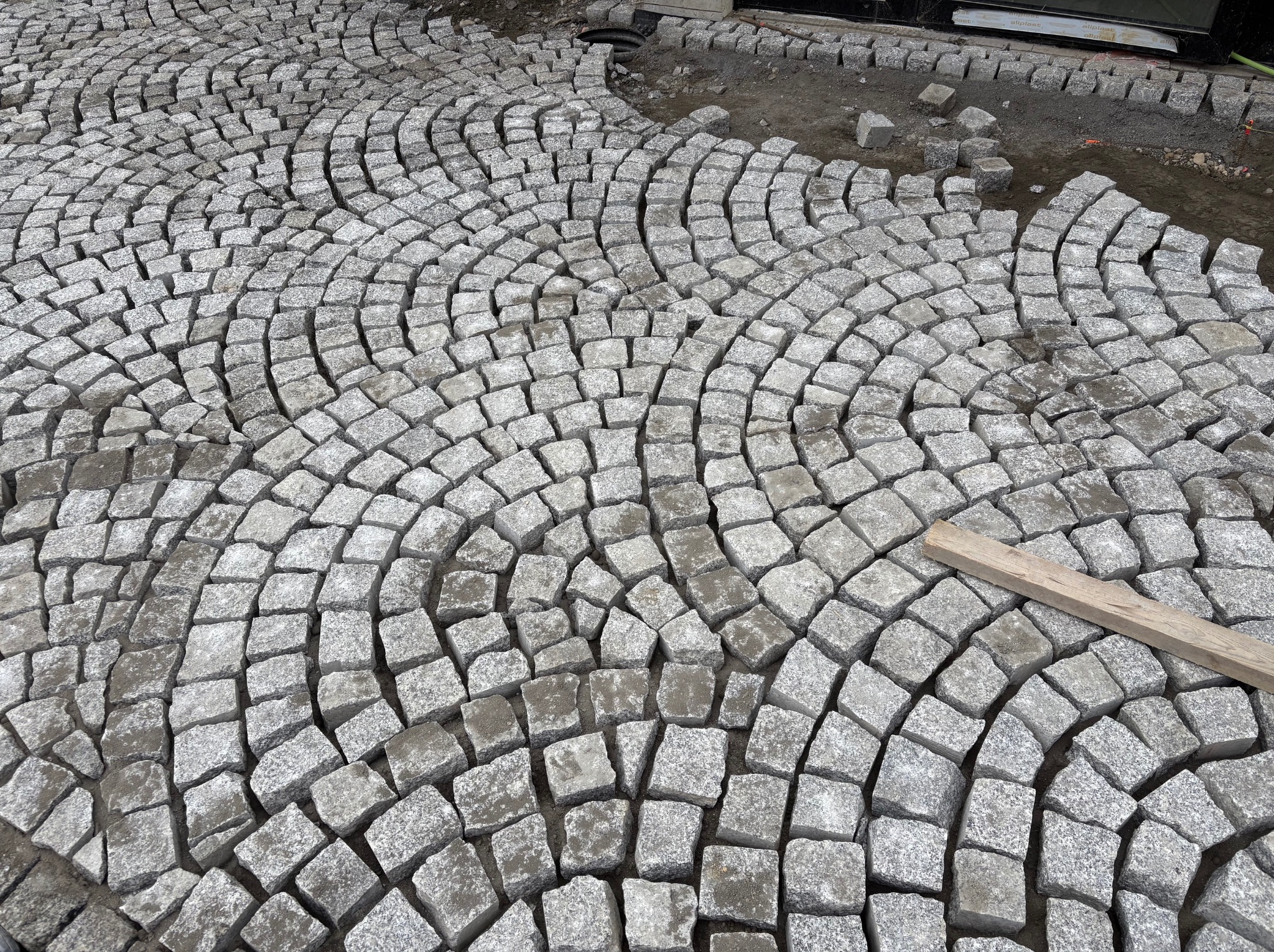 Fragment układania kostki granitowej w łukowaty wzór. Częściowo ułożona nawierzchnia z szarych kostek, widoczna drewniana listwa i niezakończony fragment.