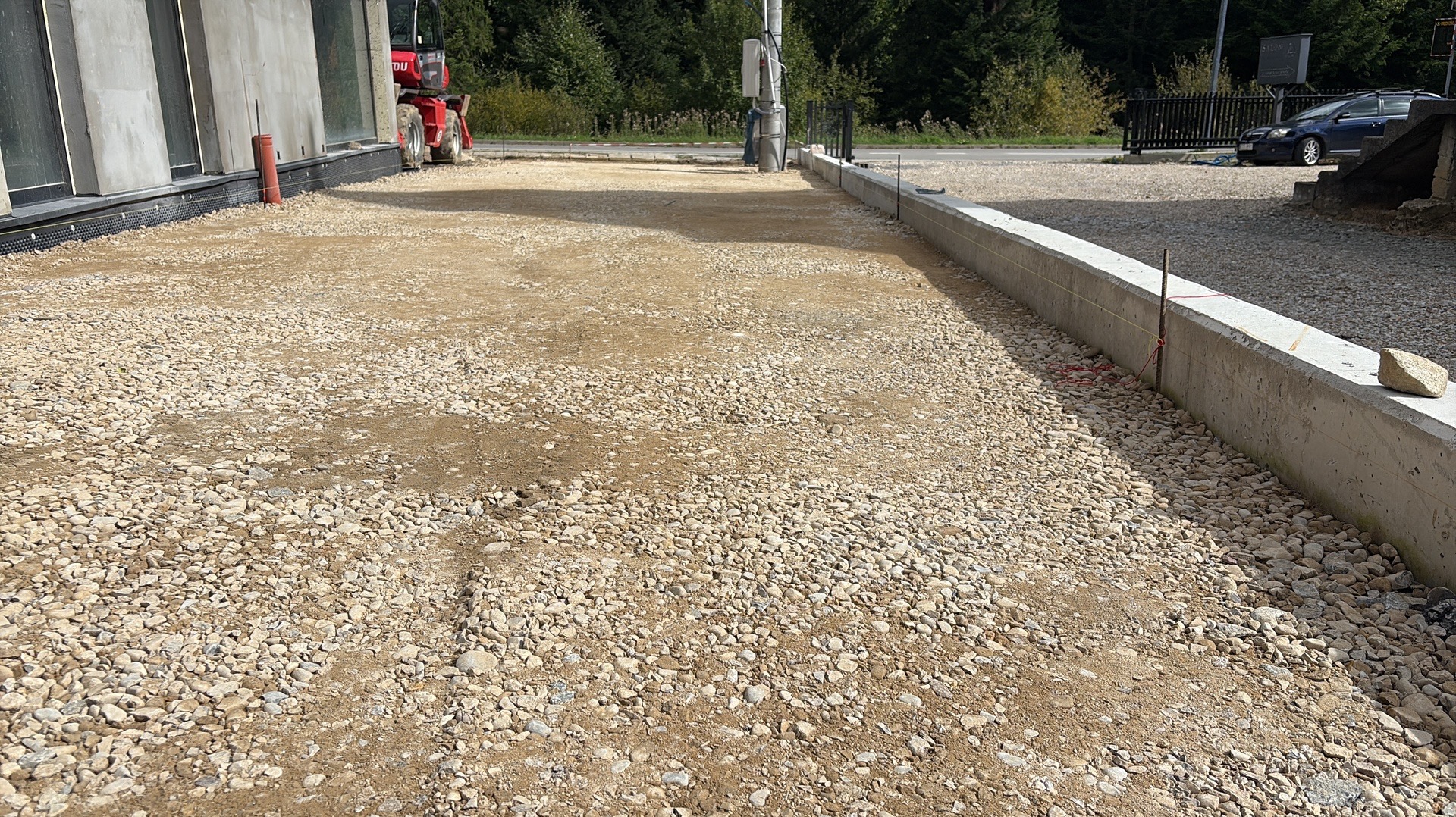 Teren przygotowany pod kostkę brukową z widocznym obrzeżem betonowym i miniładowarką w tle, ukazujący etap prac ziemnych.