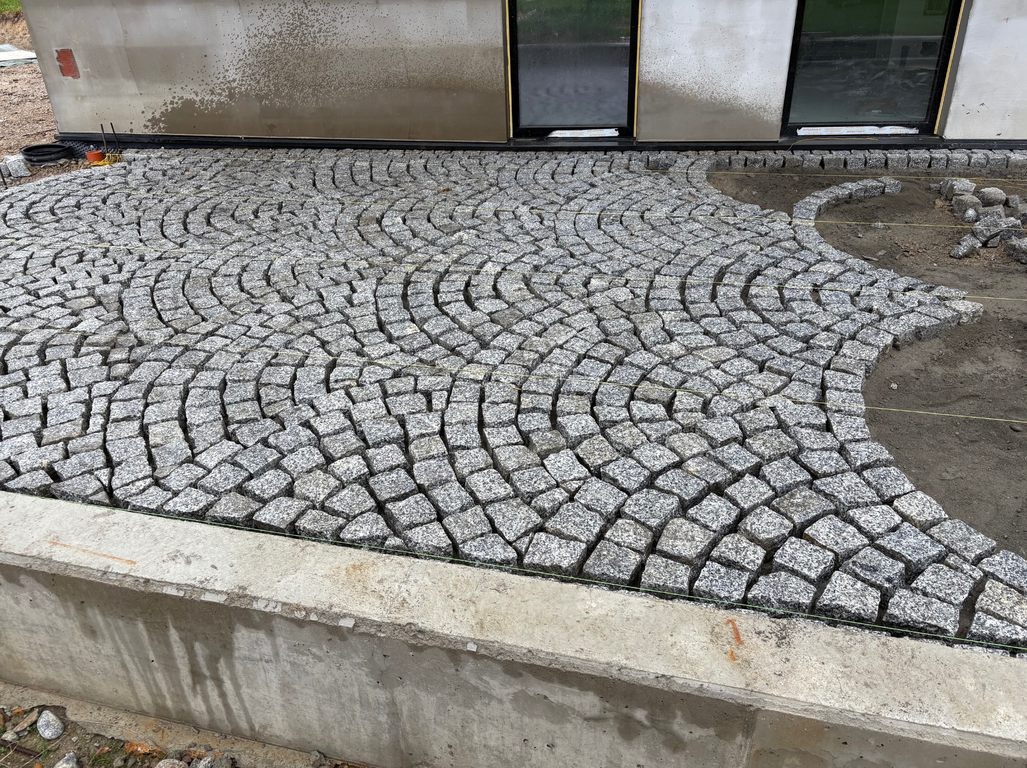 Układanie kostki granitowej w łukowaty wzór przy budynku. Częściowo ułożona nawierzchnia z kostki, widoczne sznurki wyznaczające obszar robót.