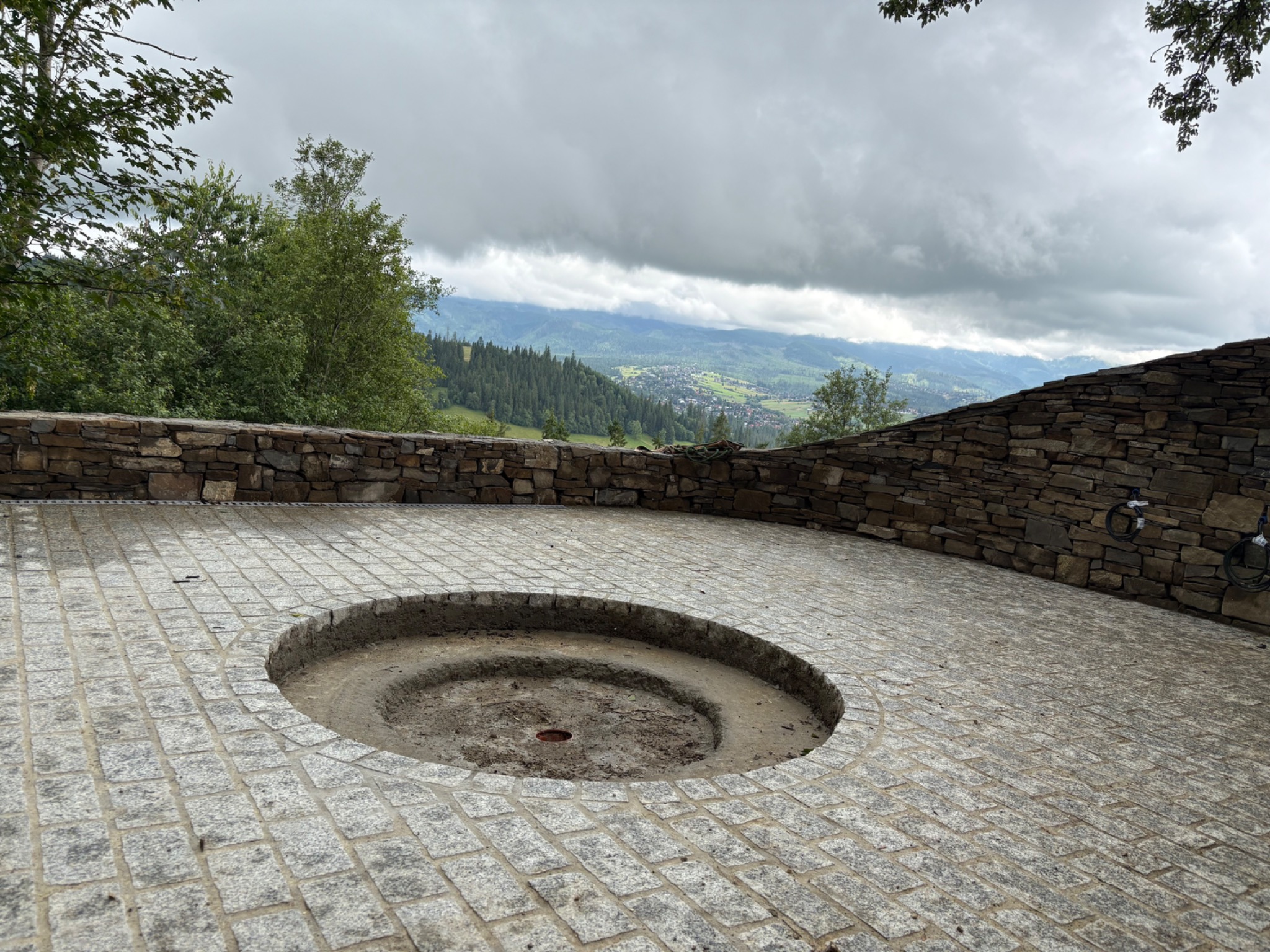 Granitowa nawierzchnia tarasu z okrągłym wgłębieniem na palenisko, otoczona kamiennym murem z widokiem na góry w tle. Pochmurne niebo.
