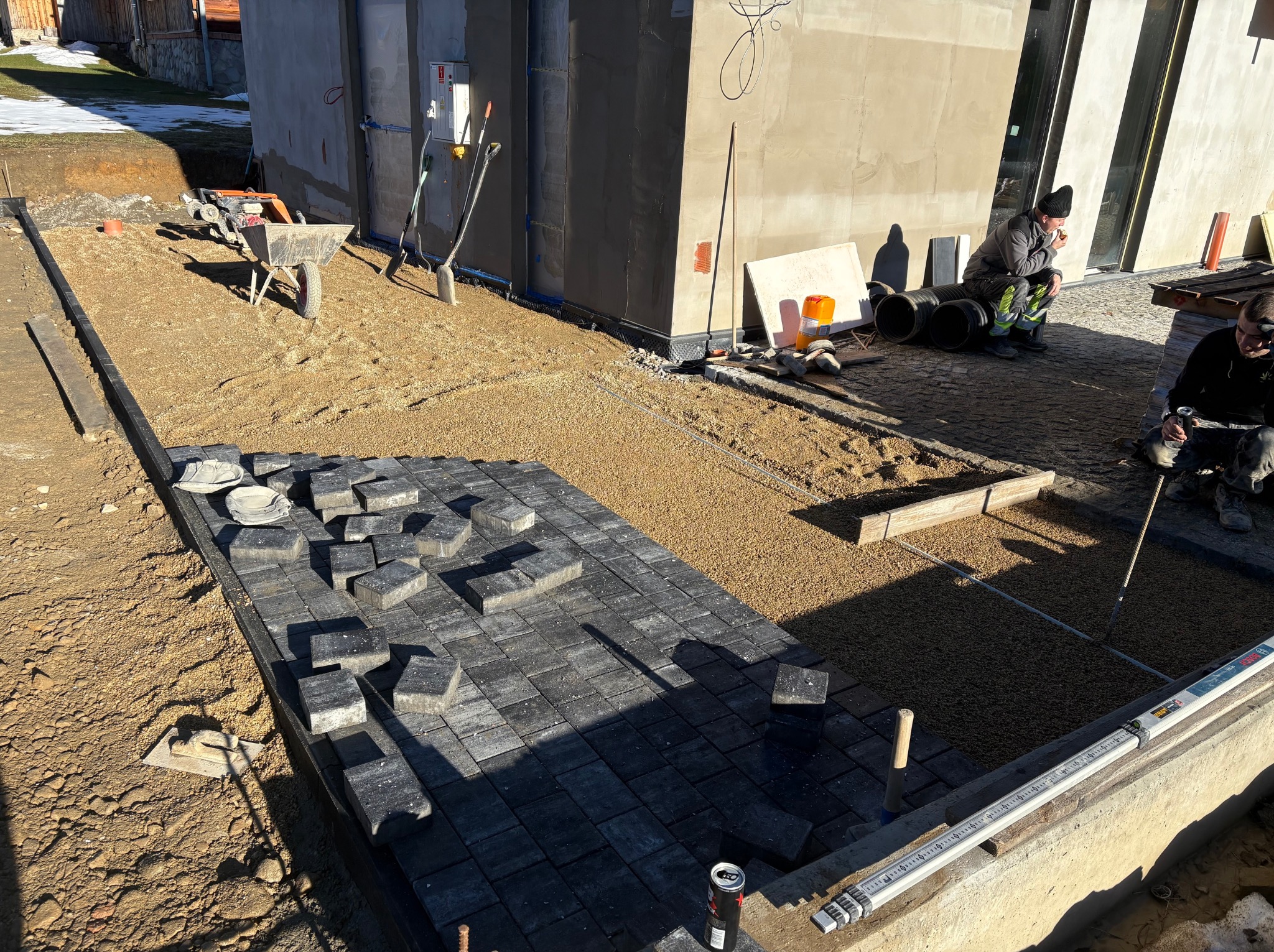 Fragment układanej, szarej kostki brukowej na podsypce piaskowej. W tle taczka, ubijarka i narzędzia. Dwóch pracowników w trakcie przerwy. Widok z góry, słoneczny dzień.