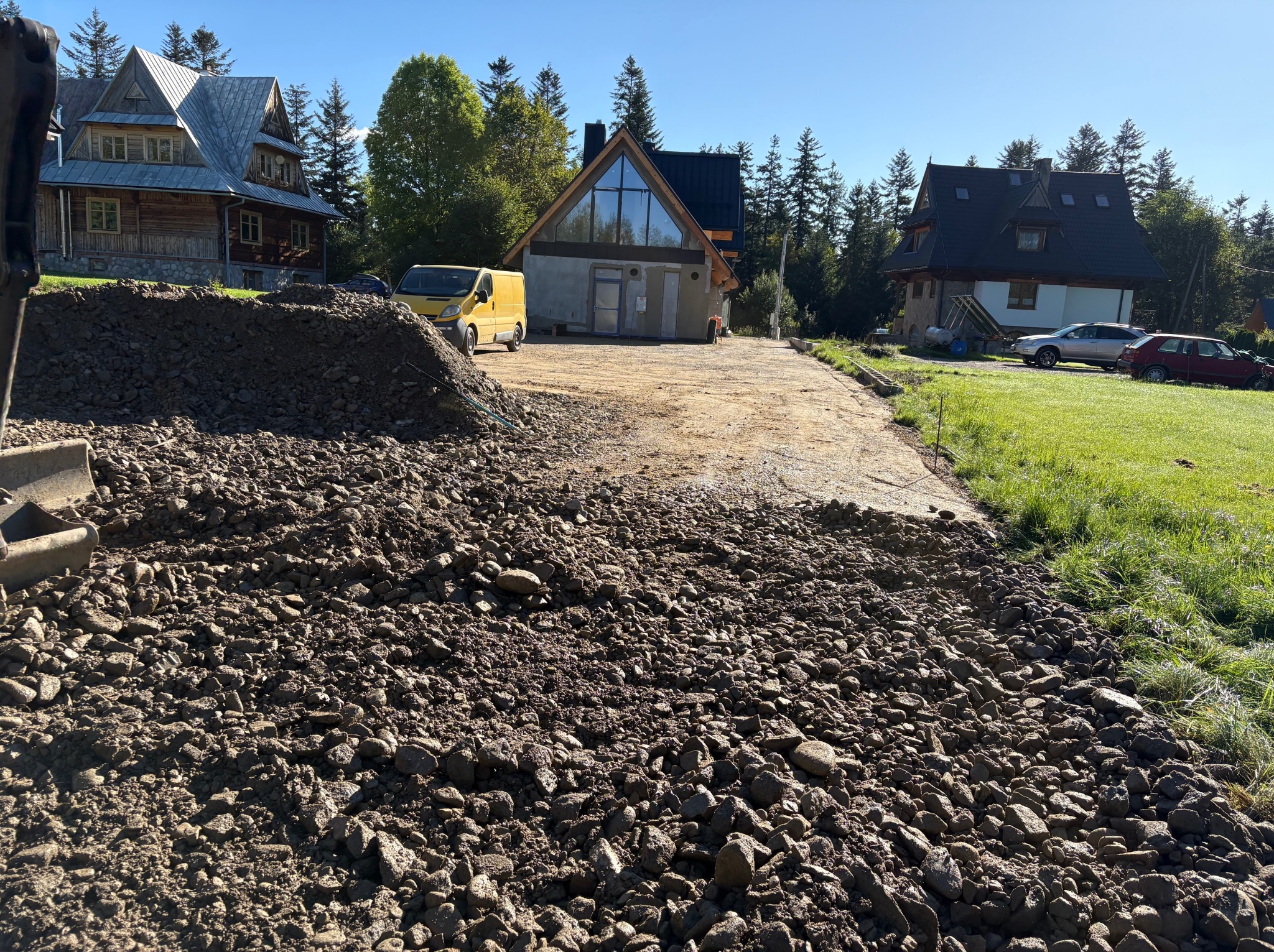 Przygotowanie podłoża pod budowę drogi dojazdowej: wysypany gruz, widoczny fragment koparki, w tle domy w stylu góralskim i żółty bus. Słoneczny dzień.