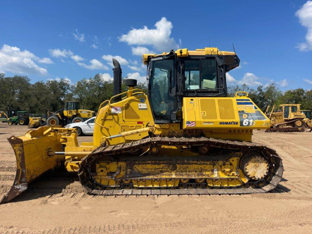 Żółty buldożer Komatsu D61 z widocznymi śladami użytkowania na gąsienicach, stojący na piaszczystym terenie w słoneczny dzień, z innymi maszynami budowlanymi w tle.
