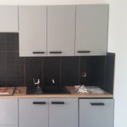 Szare szafki kuchenne z czarnymi uchwytami i czarną armaturą na tle czarnych płytek, widoczne narzędzia i akcesoria montażowe, gniazdko elektryczne w ścianie.