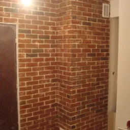 Ściana w stylu cegły w Łodzi, z widoczną kratką wentylacyjną i fragmentem drewnianej podłogi, oświetlona górnym światłem.