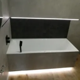 Nowoczesna łazienka z podświetlaną wanną obudowaną panelami w odcieniach szarości, czarne płytki nad wanną i drewnopodobna podłoga z oświetleniem LED.