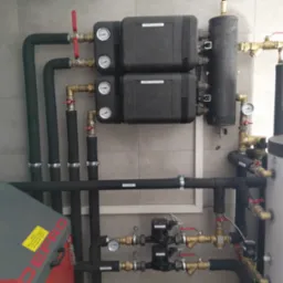 Instalacja grzewcza z dwoma modułami pompy obiegowej (grzejnikowa i podłogowa), zbiornikiem akumulacyjnym, orurowaniem z izolacją, zaworami i manometrami, na tle szarej ściany z płytek.